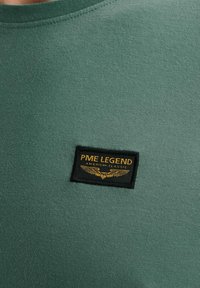 T-shirt verde in cotone con un primo piano di un badge nero che presenta la scritta "PME LEGEND AMERICAN CLASSIC" in oro, tessuto texturizzato.