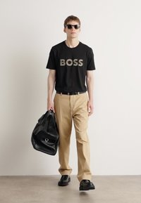 Melnas T-krekls ar "BOSS" logo, bēšas bikses, melnas kurpes, nēsā melnu sporta somu. Rūpīgi pieguļošs dizains, ikdienas stils.