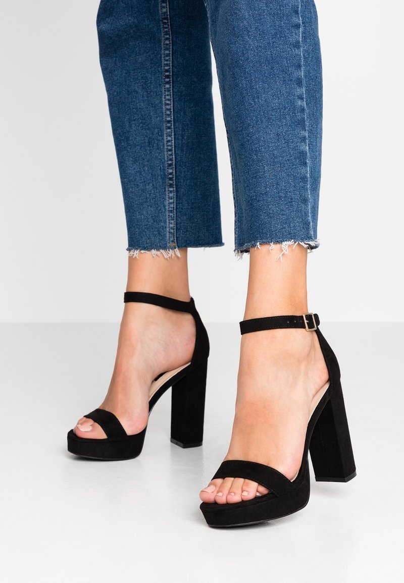 Jalkaterät mustissa korokkeet sandaaleissa nilkkatuilla, yhdistettynä vaaleisiin farkkihin, joiden helmassa on risaiset reunat.