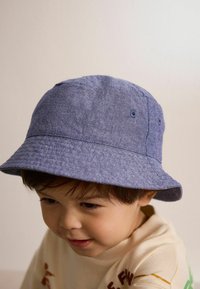 Cappello a secchiello in denim blu con superficie testurizzata, caratterizzato da due occhielli sui lati e una tesa ampia che si inclina verso il basso.