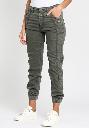 Slank pasform olivengrønne bukser med elastiske manchetter båret med hvide sneakers og en hvid top, én hånd i lommen, stående på en ensfarvet baggrund.