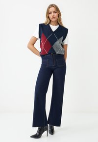 Gilet lavorato a maglia blu navale con motivo argyle rosso, grigio e bianco, indossato sopra una maglietta bianca; abbinato a jeans scuri a gamba larga e stivali neri con tacco.