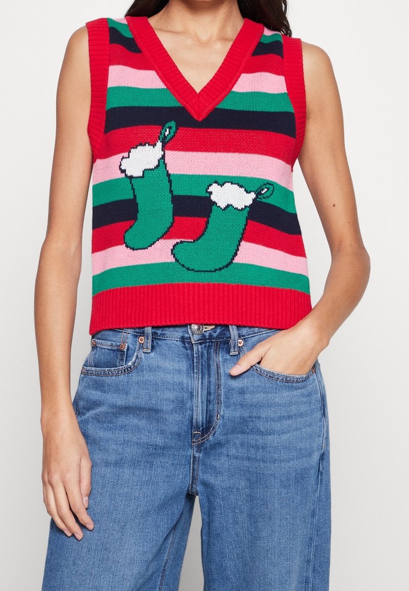Femme portant un gilet sans manches rayé rouge, rose, vert et bleu marine avec deux motifs de chaussettes de Noël vertes, associé à un jean bleu.