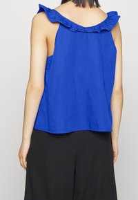 Haut bleu sans manches en tissu léger, avec un col à volants et une coupe ample, accompagné d'un pantalon sombre texturé.