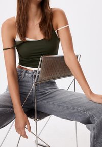 Jeune femme aux longs cheveux assise à l'envers sur une chaise en métal, portant un haut court vert foncé à fines bretelles et un jean taille haute gris.