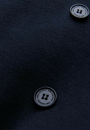 Tissu texturé bleu marine avec un bouton métallique rond comportant deux trous et du texte en relief. Le bouton contraste avec le matériau doux.