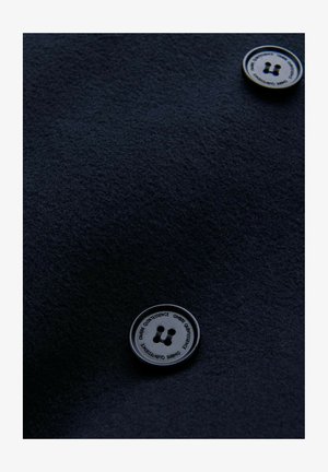 Navy textiel met een ronde, metalen knop met twee gaten en reliëftekst. De knop vormt een contrast met het zachte materiaal.