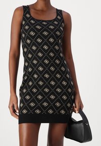 Femme portant une robe noire sans manches ajustée avec un motif de diamants beiges et de logos, tenant un petit sac à main noir à son côté.