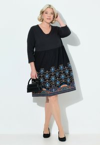 Schwarzes Kleid mit drei Viertel Ärmeln, V-Ausschnitt, gerafftem Oberteil und buntem, gemustertem Saum mit blauen Rautenmuster und floralen Akzenten.