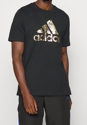 Homme portant un t-shirt Adidas noir avec un logo marbré doré et un pantalon Adidas noir avec des bandes bleues, debout devant un fond uni.