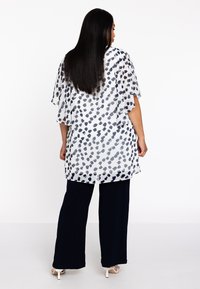Durchsichtige weiße Bluse mit schwarzen abstrakten Mustern, weiten Ärmeln, kombiniert mit marineblauen Hosen und hellen Heels. Sauberes Design, fließende Passform.