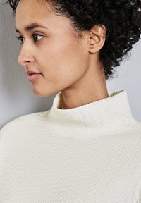 Weißer strukturierter Rollkragenpullover mit einem gerippten, erhabenen Muster. Der Kragen ist weit, und der Stoff wirkt weich und strukturiert.