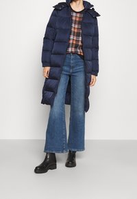 Marineblå pufferjakke med hætte, der har et quiltet design, frontlynlås og lange ærmer. Parret med en ternet sweater og vide denimjeans.