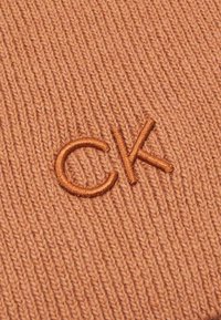 Tissu tricoté de couleur pêche chaude avec un logo brodé "CK" en pêche plus foncé. La texture est côtelée, avec une apparence douce et légèrement duveteuse.