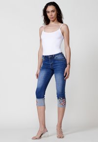 Jean capri en denim bleu avec broderie florale à l'ourlet, associé à un débardeur blanc ajusté et des sandales à talons bloc transparent.