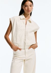 Vest alb scurt, cu fermoar, mâneci scurte și modele brodate în spirale; asortat cu pantaloni cu talie înaltă, la fel.