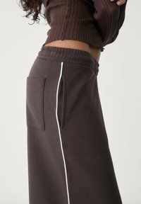 Bruine sweatpants met een gestructureerd oppervlak, voorzien van een zijzak en een witte verticale streep. Elastische tailleband voor comfort.