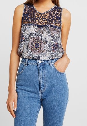 Vrouw draagt een mouwloze top met bloemenpatroon, een marineblauwe haakwerk inzetstuk op de bovenkant en een high-waisted blauwe spijkerbroek, met haar hand in de zak.