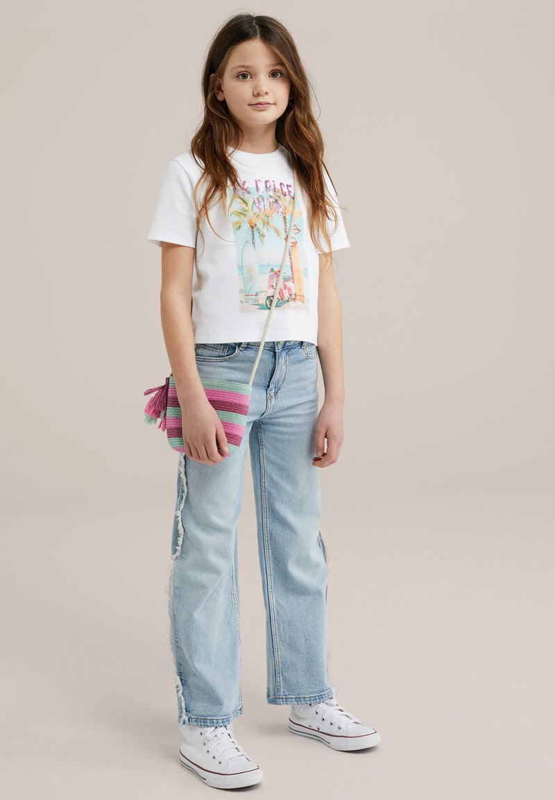 Witte grafische t-shirt met kleurrijke surf-thema print, lichtblauwe jeans met zijdelingse slijtage, en een kleine multikleurige gestreepte schoudertas.