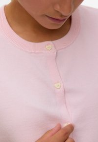 Ljusrosa stickad cardigan med rund halsringning, som har två hjärtformade knappar och ribbat mönster längs kragen och knapplisten.
