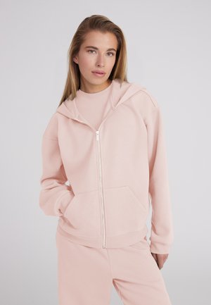 Junge Frau mit langen blonden Haaren, die eine hellrosa Zip-Hoodie und passende Hose vor einem einfarbigen grauen Hintergrund trägt.