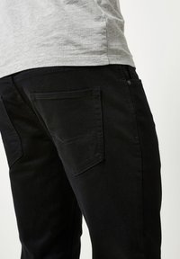 Mustat farkut slim fit -mallilla, joissa on kaksi takataskua ja hienovaraisia ompeluyksityiskohtia, harmaan t-paidan taustalla.