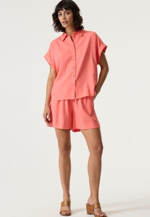Camicia a maniche corte color corallo con bottoni abbinata a pantaloncini dello stesso colore, caratterizzata da una vestibilità comoda e tessuto morbido, completata da sandali con tacco color cuoio.