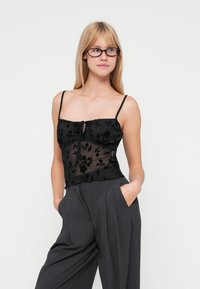 Camisola nera in pizzo floreale con spalline regolabili, caratterizzata da un dettaglio con nodo anteriore e sovrapposizione trasparente, abbinata a pantaloni grigi a pieghe larghi.
