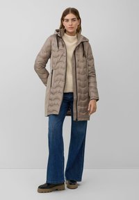 Abrigo acolchado de color marrón claro con capucha, cierre de cremallera y bolsillos laterales, combinado con unos vaqueros de pierna ancha de color azul y botas de plataforma negras.