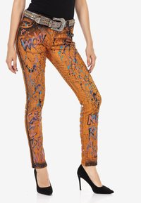 Jambes vêtues d'un pantalon orange éclaboussé de graffiti avec des mots, ceinture ornée de strass, haut noir et chaussures à talons noirs sur fond blanc.