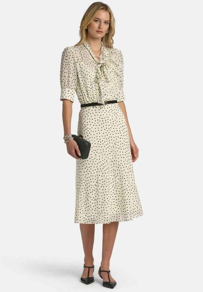 Krém színű pöttyös ruha fekete pöttyökkel és masnis gallérral. Rövid, buggyos ujjakkal, deréknál megkötve fekete övvel, és lebegő szoknyával.