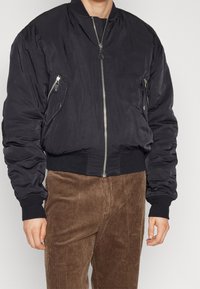 Schwarze Bomberjacke mit Reißverschlusstaschen, gerippten Bündchen und Saum. Glatte Textur und gepolstertes Design. Kombiniert mit braunen Cordhosen.