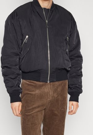 Giacca bomber nera con tasche con zip, polsini e vita a coste. Texture liscia e design imbottito. Abbinata a pantaloni in velluto a spiga marroni.