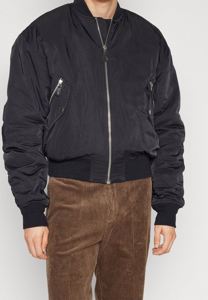 Schwarze Bomberjacke mit Reißverschlusstaschen, gerippten Bündchen und Saum. Glatte Textur und gepolstertes Design. Kombiniert mit braunen Cordhosen.