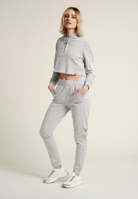 Haut hoodie gris court et joggeurs assortis en matériau doux avec poches. Détails du logo et motif en chevron sur les joggeurs, associés à des baskets blanches.