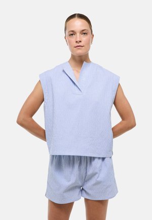 Femme à la peau claire et aux cheveux tirés en arrière porte un haut sans manches ample à rayures bleues et blanches et un short assorti, debout avec les mains dans le dos.