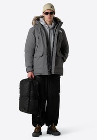 Grå isolerad jacka med pälsfodrad huva, grå munkjacka under, svarta byxor, bär en svart ryggsäck, har solglasögon och en beigefärgad mössa.