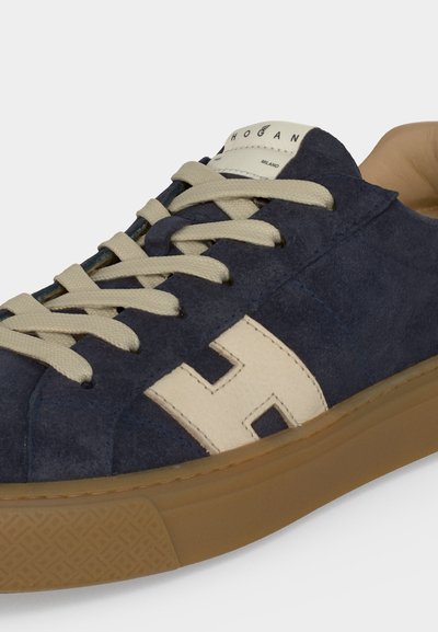 Gros plan d'une basket en daim bleu marine avec des lacets beige, un logo blanc "H" sur le côté et une semelle en caoutchouc marron clair.