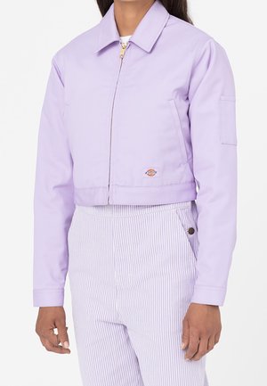 Kvinde iført en lyse lilla cropped jakke med frontlynlås og højtaljede bukser med striber, stående mod en hvid baggrund.