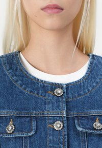 Denim jakna s srebrnimi cvetličnimi gumbi, modra tkanina, krožni izrez in bela srajca vidna spodaj. Prisoten je ključni šivni detajl.
