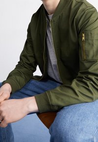 Giacca bomber verde oliva con zip frontale, polsini a coste e tasca con zip sulla manica, indossata sopra una t-shirt grigia e jeans blu. Texture liscia.