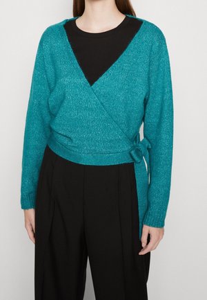 Femme portant un pull cache-cœur turquoise noué sur le côté par-dessus un haut noir et un pantalon noir ample, debout devant un fond uni.
