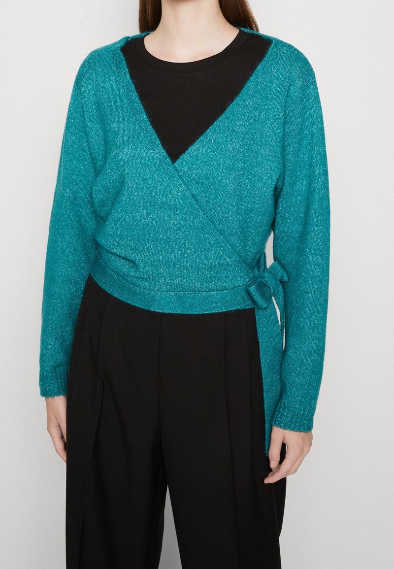Femme portant un pull cache-cœur turquoise noué sur le côté par-dessus un haut noir et un pantalon noir ample, debout devant un fond uni.