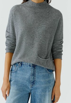 Grå langærmet strikket trøje med lille frontlomme, båret med lyseblå højttaljede jeans, ærmerne skubbet op til underarmene.