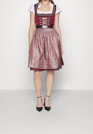 Kobieta ubrana w burgundową sukienkę dirndl z białą falbaniastrą bluzką, metalicznie różowym fartuchem i czarnymi szpilkami na kostkę, stojąca na białym tle.