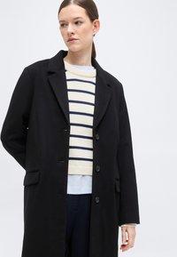 Γυναίκα με σκουρόχρωμα μαλλιά που φοράει μαύρο παλτό με κουμπιά πάνω από λευκό πουλόβερ με ναυτικούς οριζόντιες ρίγες, κοιτάει προς τα αριστερά της σε απλό φόντο.