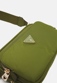 Borsa a tracolla verde oliva con zip dorata e logo triangolare Guess sul davanti, dotata di tracolla in tessuto regolabile.