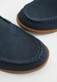 Mørkeblå ruskinds loafers med kontrastfarvet brun gummisål, prydet med dekorativ syning langs kanterne og en glat tekstur.