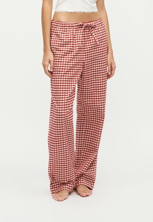 Femme portant un pantalon ample à carreaux rouges et blancs et un haut court blanc, debout pieds nus sur un fond blanc uni.