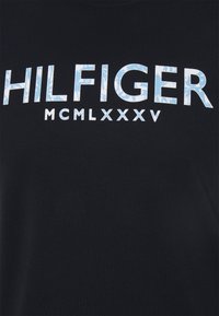 Mustat kangas, jossa on suuret siniset ja valkoiset marmoroidut kirjaimet "HILFIGER" ja pienemmät valkoiset tekstit "MCMLXXXV" alla.